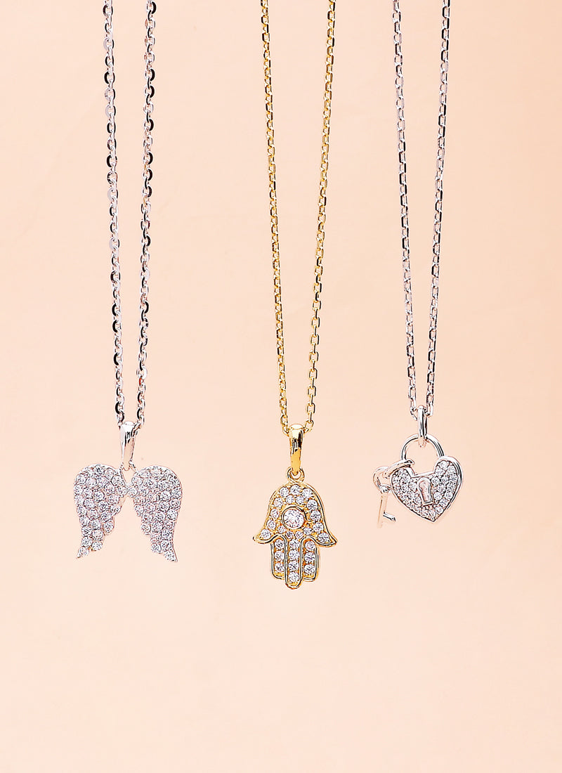 Three sparkling pendant necklaces featuring symbolic designs: an angel wing, a Hamsa hand, and a heart-shaped lock with a key, displayed on delicate chains against a soft beige background.