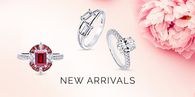 New arrivals featuring a bold red gemstone ring, a modern baguette diamond ring, and a classic round-cut solitaire ring, set against a soft pink background with delicate flowers.